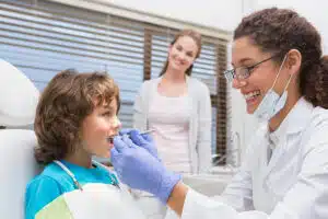 A smiling female dentist examining a young boy's teeth while his mother watches in the background.