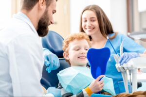 A young boy smiling while looking at his teeth in a blue tooth-shaped mirror at the dentist